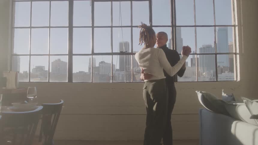 Couple dances together in sunlit room with city view through the windows