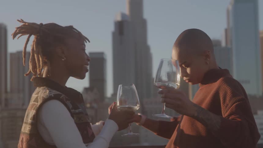 A young black couple dancing together while holding drinks on a balcony in a metropolitan city during day