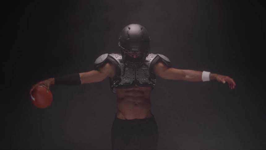 A rugby player wearing protective gear and a helmet holds a football with arms spread apart