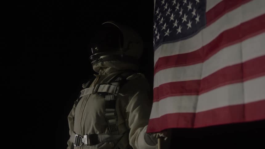 An astronaut in a spacesuit stands proudly next to a large american flag against a completely black background