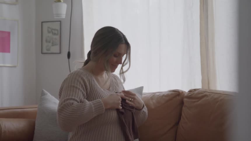 A pregnant woman wearing a ribbed beige sweater and skirt sits on a brown leather sofa in a bright living room and looks at a small piece of dark baby clothing