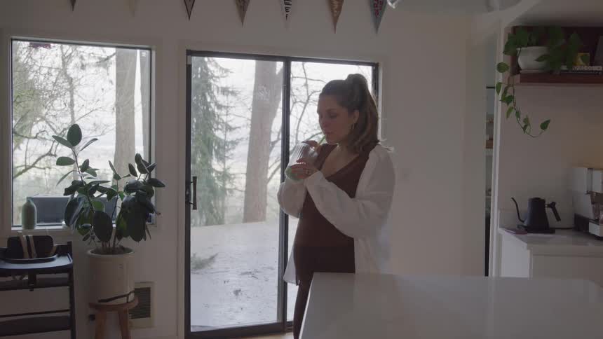 A pregnant woman with long hair pulled back wearing a brown top and white shirt stands next to a glass door and window in a kitchen stirring and drinking a beverage