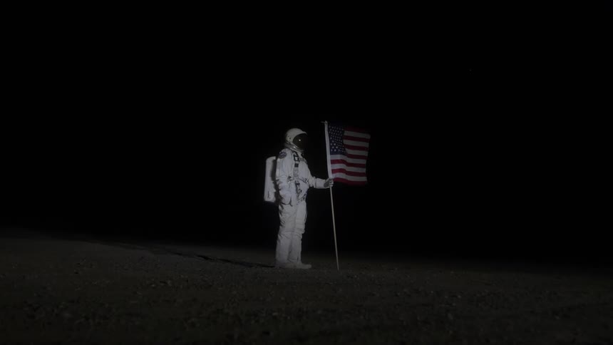 An astronaut stands on a barren surface holding the american flag