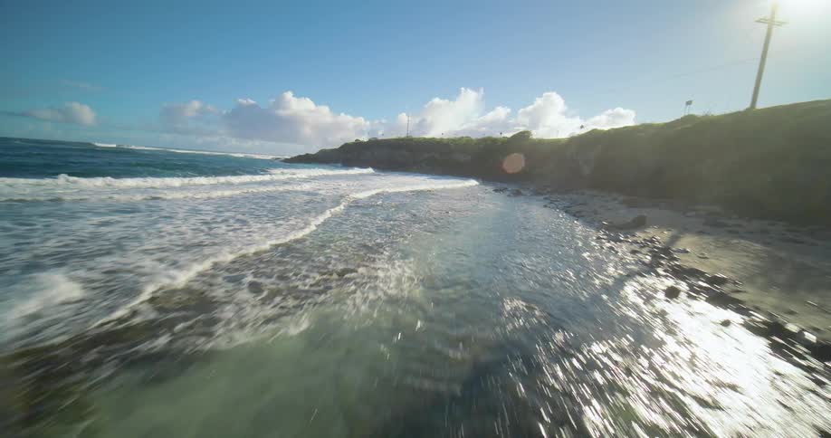 An aerial low view glides rapidly over a sandy beach and rocky shallow water with ocean waves crashing toward a road lining the green coastal cliffs under a bright blue cloudy sky