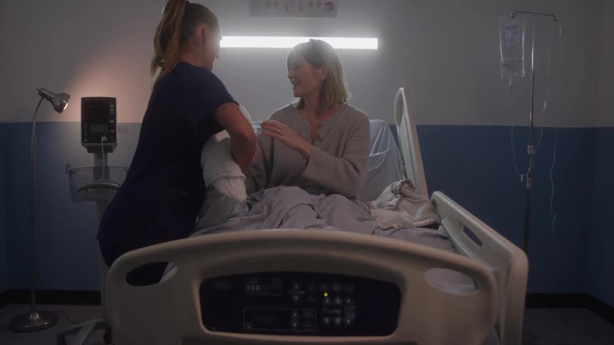 A nurse wearing dark blue scrubs assists a woman wearing a gray shirt who is sitting up in a hospital bed with an iv bag and medical equipment visible in the dimly lit hospital room
