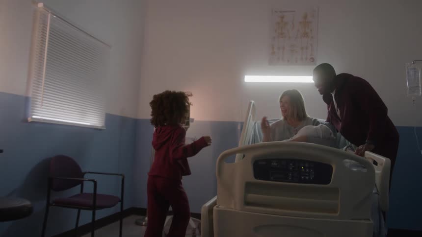 A young girl enters a hospital room and joyfully embraces her mother who is holding a newborn baby