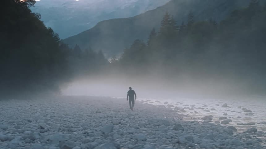 A silhouetted man walks away from the camera across a wide riverbed covered in white rocks and shrouded in thick ground fog with dark forests and mountains rising in the background