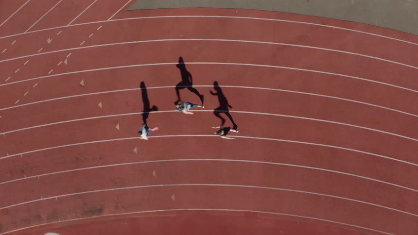 Birds eye view above three male athletes casting long shadows running while competing on an outdoor track during daytime