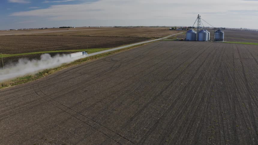 White truck kicks up dust while driving on dirt road between plowed field and grain silos