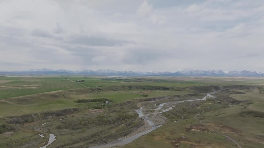 A winding river snakes through rolling brownandgreen plains toward distant snowcapped mountains under an overcast sky