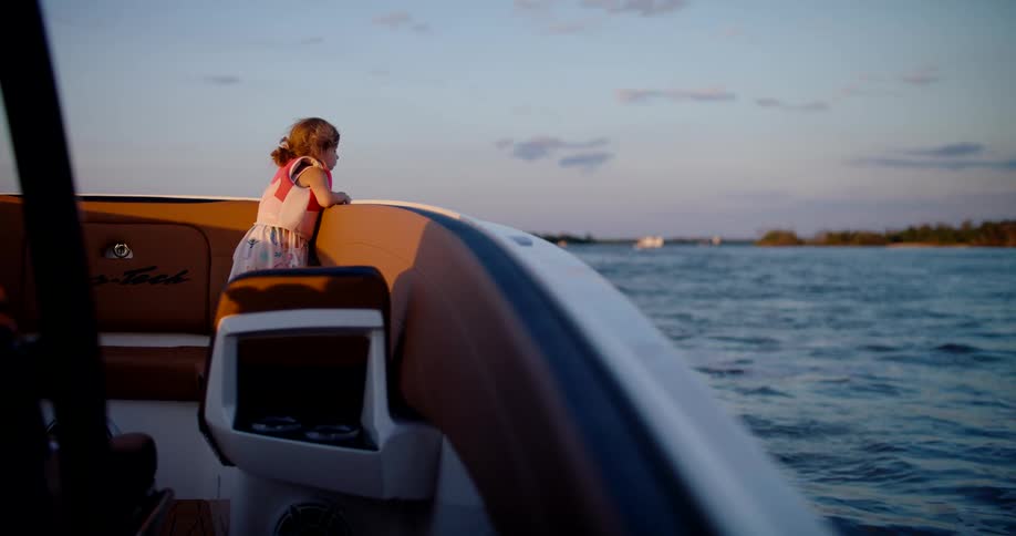 Child gazes at the water at sunset standing on boat in colorful clothes with a thoughtful expression