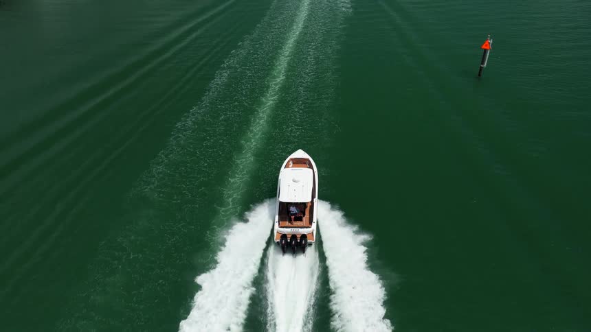Aerial view of a white motorboat moving forward across a body of green water leaving a wide prominent trail of churned water with a vertical marker visible to the right