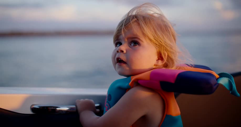 A toddler in a bright life jacket looks up wideeyed aboard a boat at golden hour wind lifting wisps of hair
