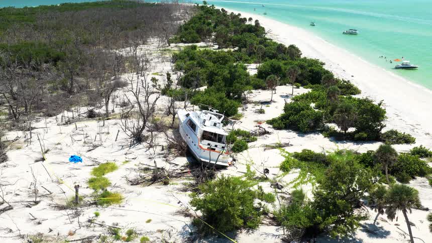 An aerial view shows a large white cabin cruiser yacht heavily beached on a white sand beach surrounded by sparse green foliage and dead trees near the turquoise ocean on a bright sunny day