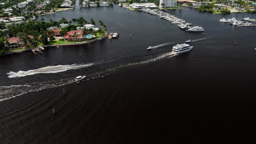 An aerial view shows a large white yacht and a small speedboat cruising down a wide dark canal past waterfront homes and a marina full of docked boats