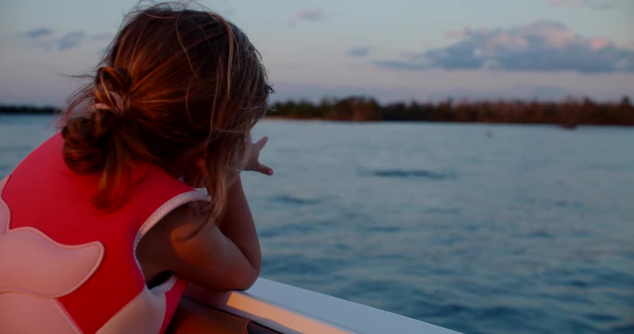Little girl wearing a life jacket smiles while leaning on the boats edge at sunset