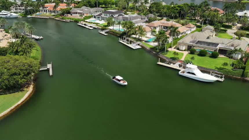 An aerial wide view shows a small white motorboat cruising through a green residential canal lined with luxurious waterfront homes docks and a large docked yacht on a sunny day