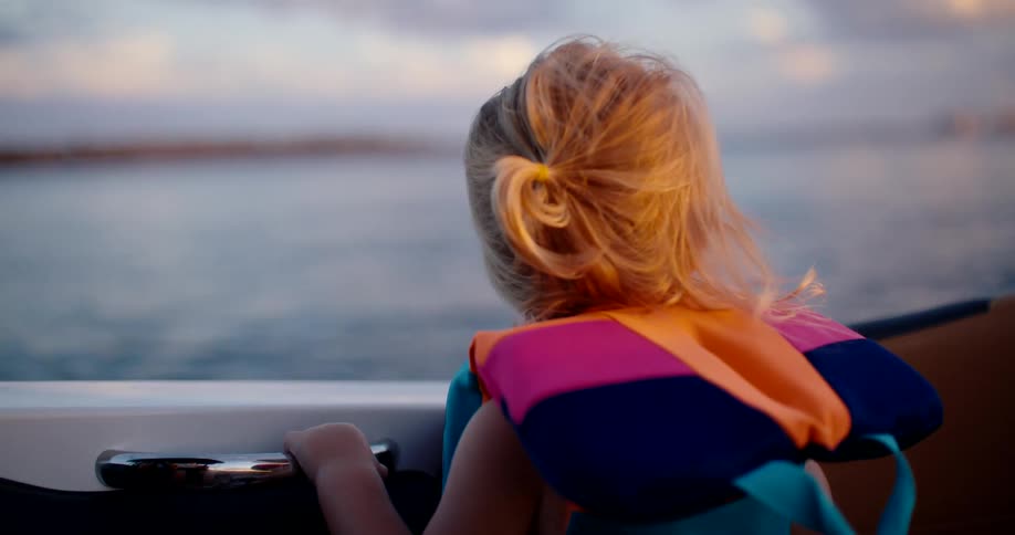 View from behind of a young girl wearing a colored flotation device gripping a handle on a watercraft and looking out over a wide body of water