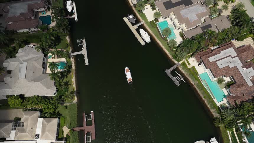 Boat sails slowly through a canal surrounded by houses with pools and green areas