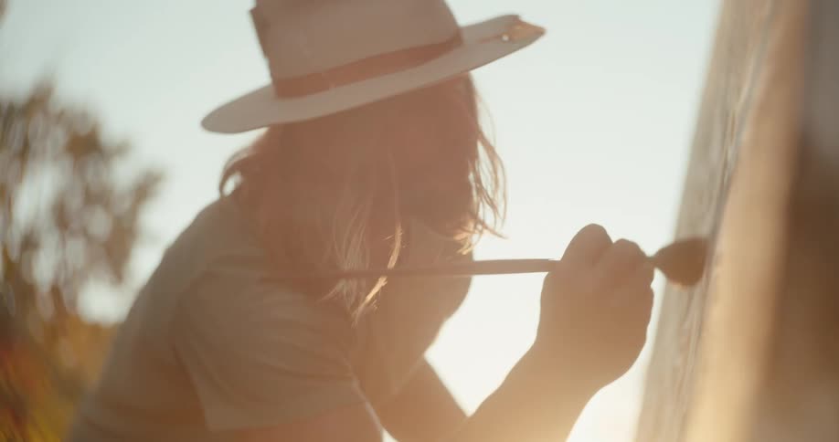 A man paints on a canvas while standing outdoors at sunset