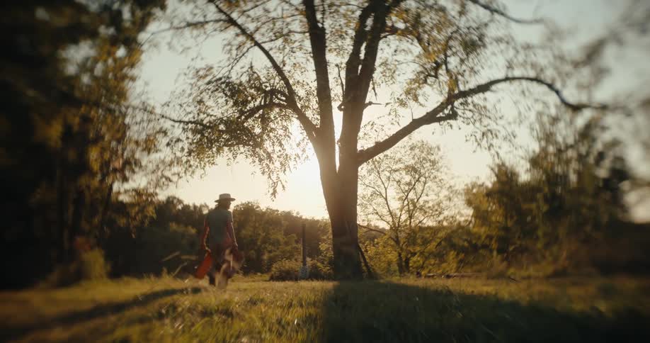 Person wearing hat carrying box across grassy field at sunset toward a tree
