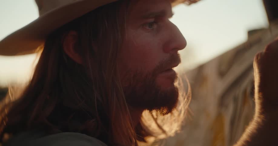 A close view shows the profile of a man with long hair and a beard wearing a cowboy hat looking intently while touching a canvas in the bright golden evening sunlight