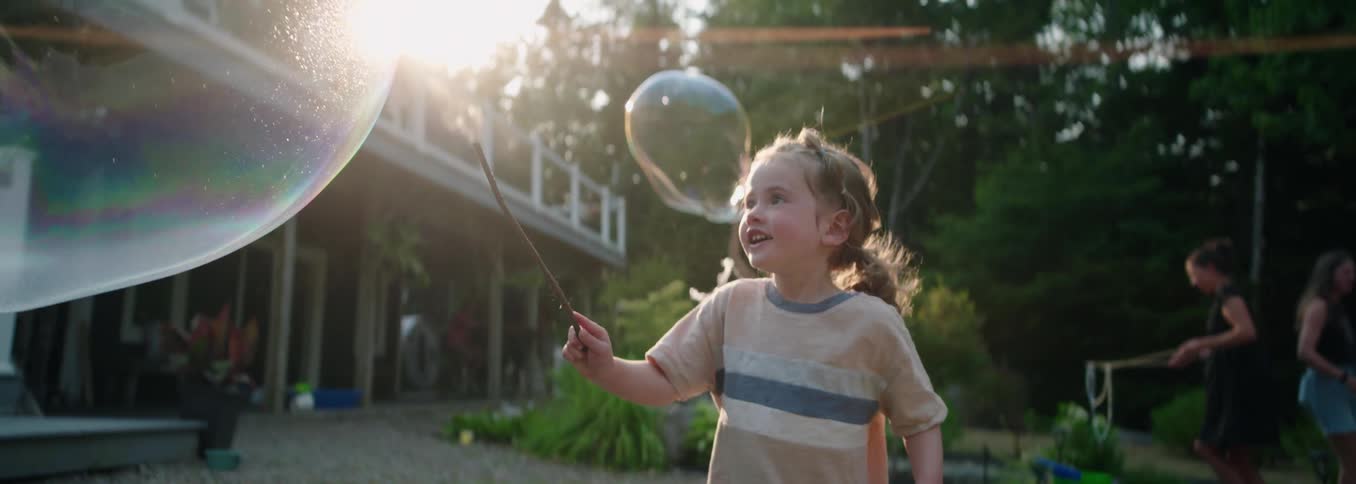 A happy young girl wearing a striped shirt watches a large soap bubble floating in the sunny backyard of a house with an adult figure blurred in the background