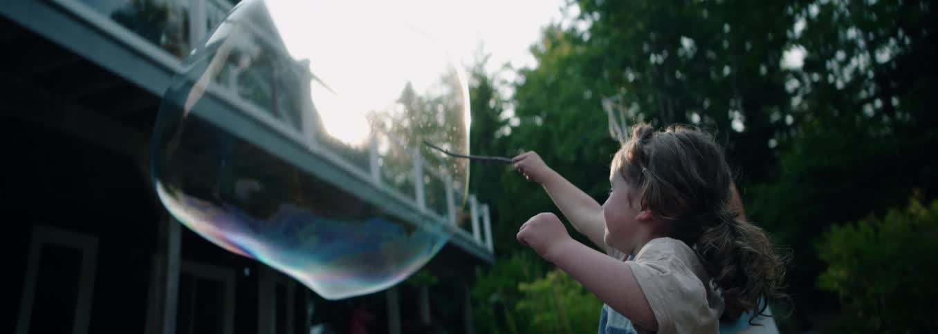A child in a backyard reaching out with a long stick to pop a large floating spherical bubble with a woman in the background