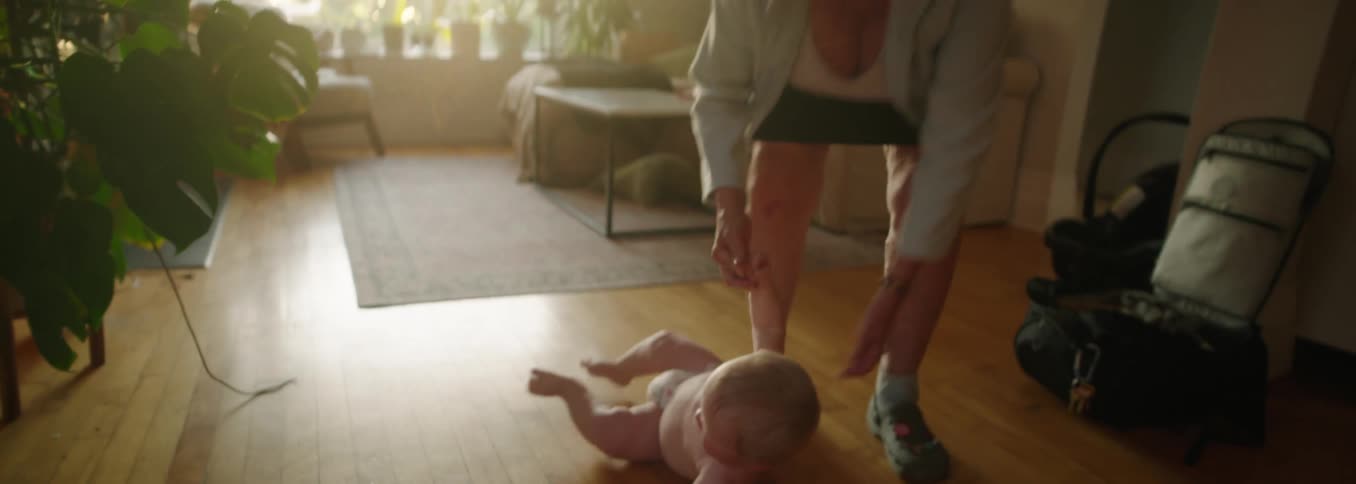 Baby takes its first steps with the support of an adult while another child watches from the couch