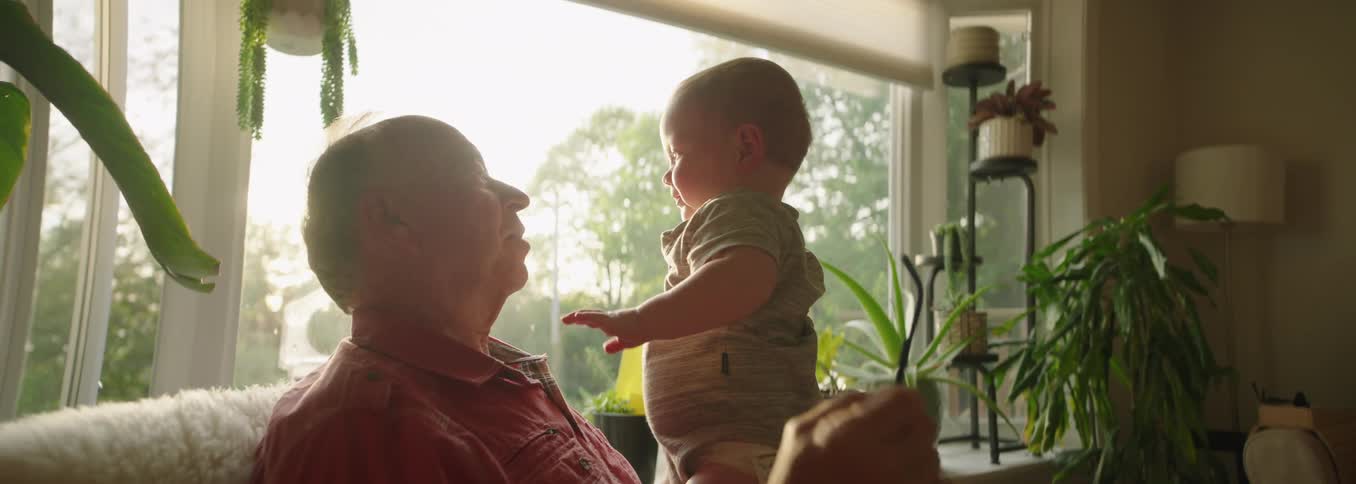 An older man wearing a red shirt and glasses holds a baby in his arms sitting near a large window with bright sunlight and houseplants creating a warm interior scene