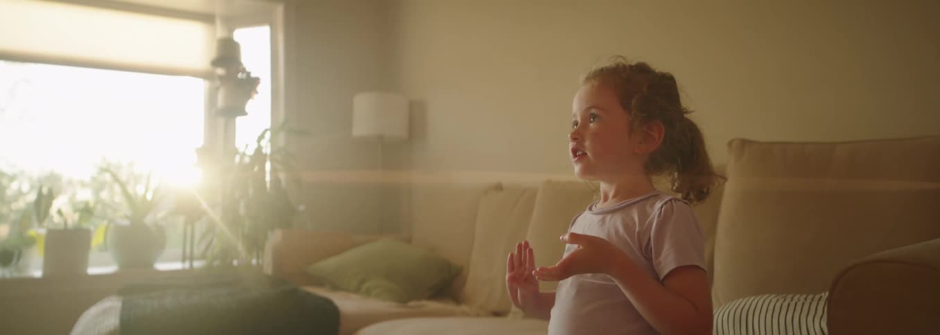 A happy young girl with brown hair in a ponytail and a light purple tshirt stands smiling and talking in a sunlit living room with a couch and houseplants visible in the background