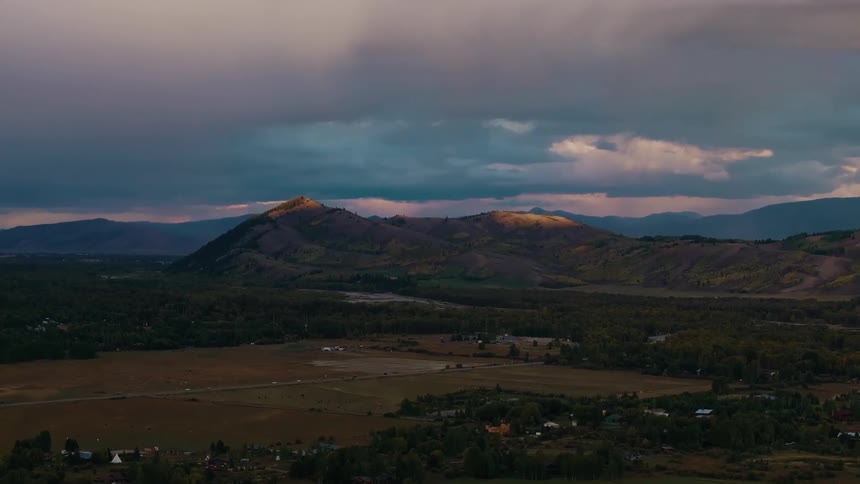 Stormy evening light brushes the tops of low mountains while a broad valley of fields and scattered homes lies below