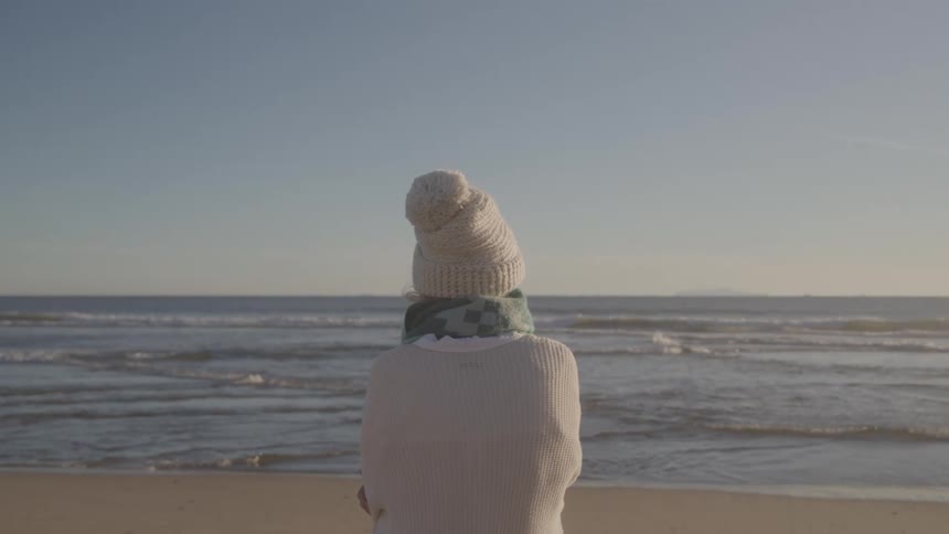 Person wearing a hat and scarf looks at the sea on a clear cold day