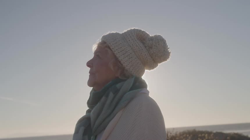 A medium side view shows an older woman wearing a white knit hat teal scarf and shawl standing on a sandy beach looking out at the ocean horizon with the bright sun creating a strong backlight