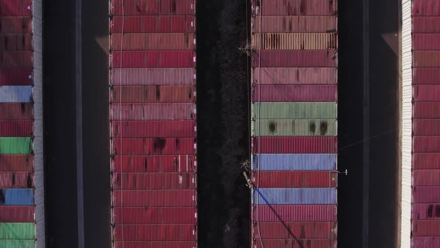Highangle aerial view slowly moving forward over multiple long straight parallel stacks of vertically arranged multicolored steel cargo boxes separated by narrow dark lanes