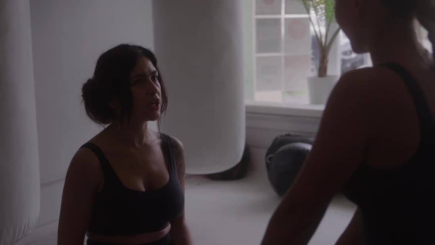 Two women in workout clothes talk inside a gym studio