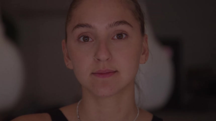 A close view shows a young womans face with her hair pulled back looking down and then smiling softly directly at the camera in a dimly lit gym with two blurred punching bags behind her