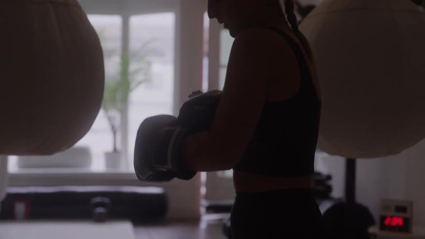 A silhouetted woman wearing a crop top and boxing gloves stands between two large heavy bags moving as if she is sparring in a dimly lit gym with a bright window in the background