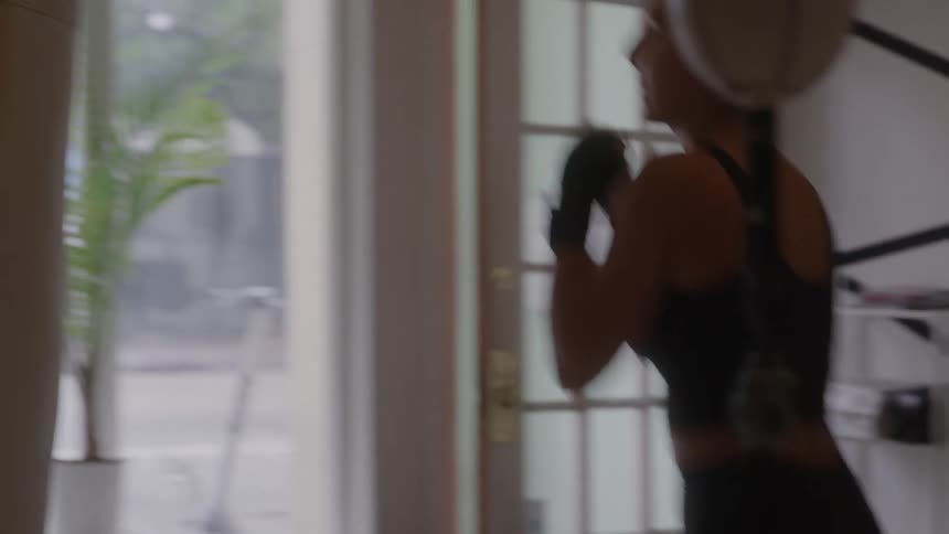 A woman performing punching movements with her hands wrapped rapidly moving her arms in rhythm with a spherical object positioned indoors near a glasspaned door