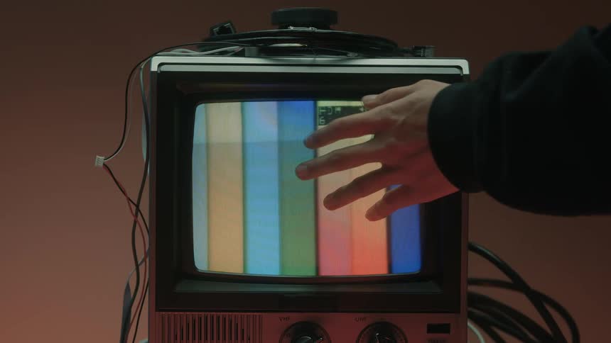 A hand reaches out and touches the screen of a vintage television which displays a classic color bar test pattern