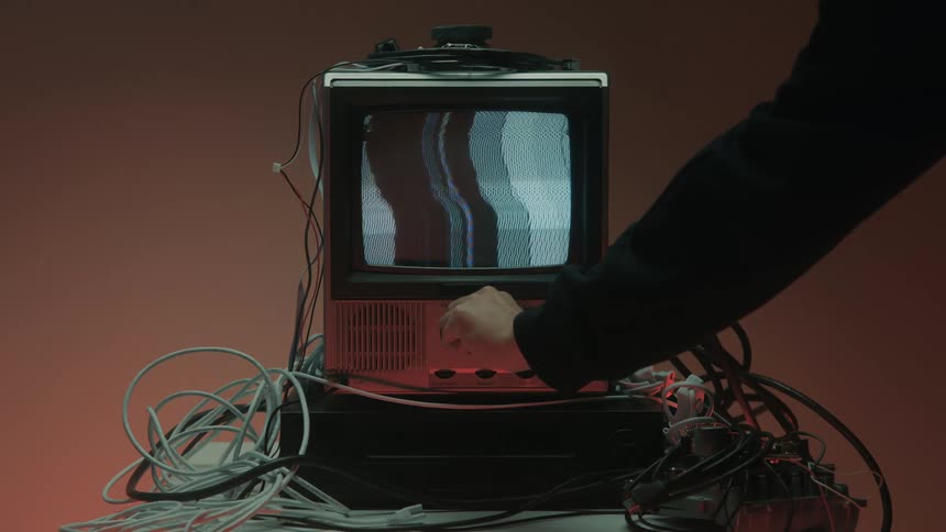 A persons arm wearing a dark sleeve adjusts a knob on a small vintage crt television with a staticfilled screen that flickers with vertical bars surrounded by a tangle of white and black cables on a burnt orange background