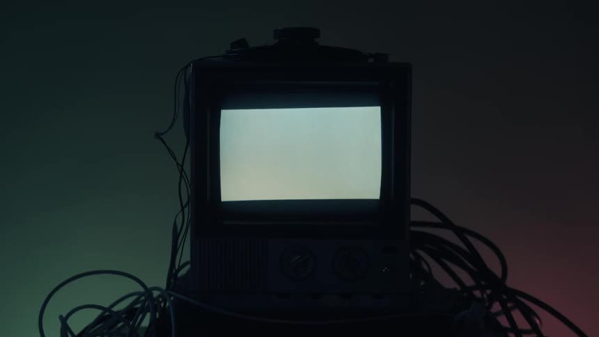 This video features a dark closeup of a vintage television or monitor surrounded by a messy tangle of cablesthe screen abruptly brightens to a solid white light for a moment then dims and reverts to a single bright horizontal line that f
