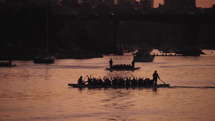 Two silhouettes of longboats likely dragon boats are visible on the water as the sun sets the larger boat in the foreground is filled with people paddling creating a striking scene against the glowing golden river