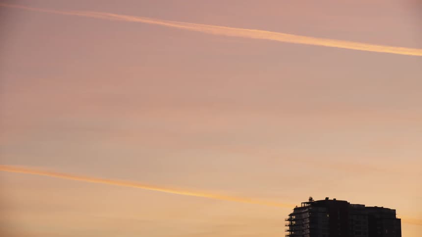 This static highangle shot captures a serene sunset or sunrise sky featuring soft hues of pink and orangetwo distinct long thin contrails vapor trails stretch diagonally across the vast expanse of the sky above the dark silhouette of