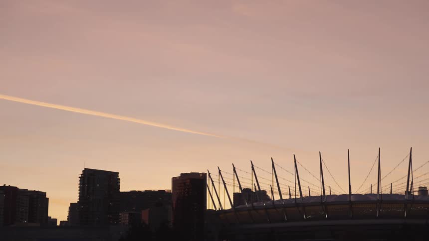 Urban skyline at sunset with a stadium and buildings beneath a pink sky crossed by an airplane trail