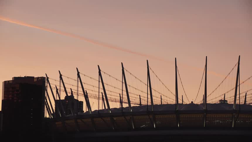 The silhouetted structure of a modern stadium and nearby buildings is set against a vibrant pale orange sky at dusk
