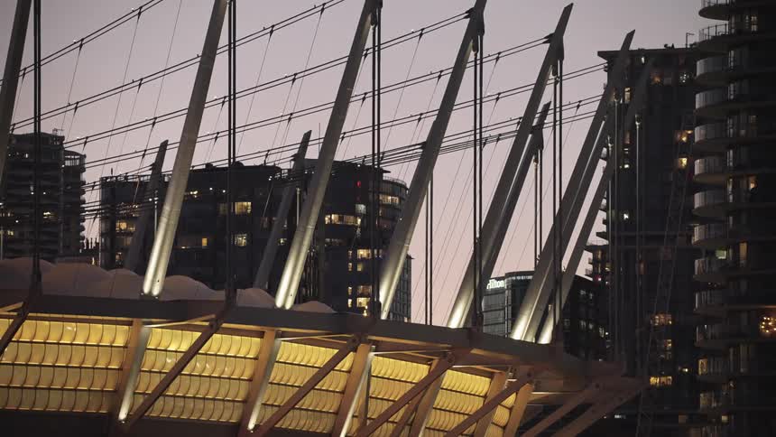 A modern stadium roof with tall spires and cable supports glows gold at dusk city towers behind