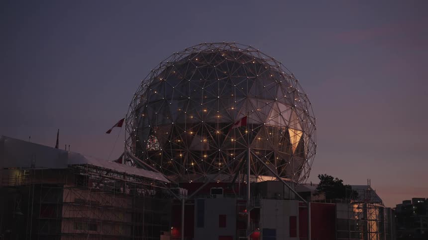 A large spherical latticestructured building with interior lights visible set against a twilight sky surrounded by lower structures with flags flying