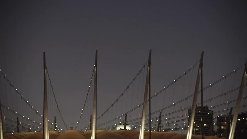 At night the illuminated architectural roof of a large arena is shown as a helicopter flies by in the distant sky