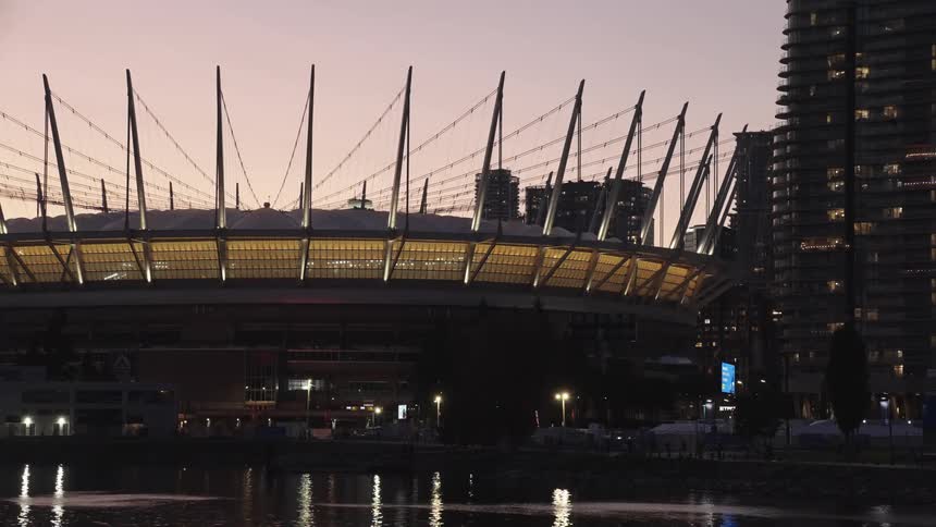 This shot taken at dusk or twilight captures the exterior of a large illuminated modern stadium with a prominent tensile roof structureit is viewed from across a body of water that reflects the lights of the stadium and the adjacent da